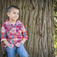 Adorable little boy in plaid shirt and jeans leaning against tree