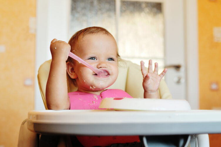 baby feeding herself cereal with spoon in highchair