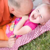 Dad tickling baby girl on blanket at the park