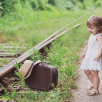 Toddler girl in sandals with suitcase by train track