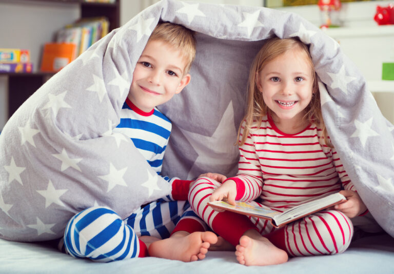 two kids reading a book in their pj\'s under a blanket