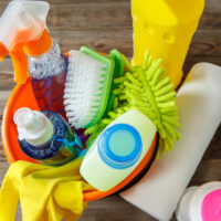 Plastic bucket with cleaning supplies on wood background. Focused on green brush.