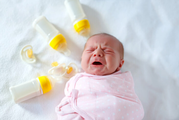 crying newborn baby girl laying on bed surrounded by bottles and pacifier