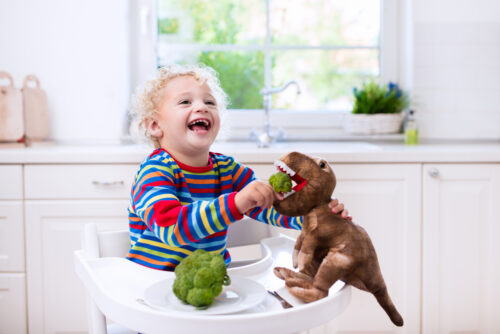 boy sitting in high chair feeding broccoli to toy dinosaur